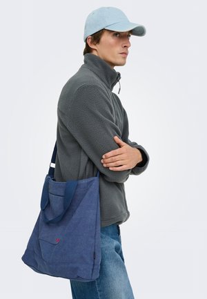 Homme avec casquette bleue et veste polaire grise, porte un grand sac bleu à l'épaule, regardant vers la droite les bras croisés.