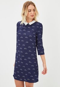 Mujer vistiendo un vestido azul marino con estampados de bicicletas blancas y un color blanco puntiagudo, de pie contra un fondo claro y liso.