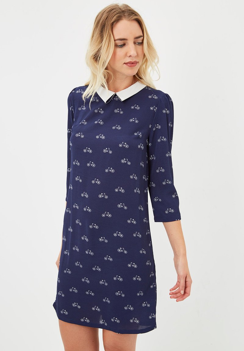 Mujer vistiendo un vestido azul marino con estampados de bicicletas blancas y un color blanco puntiagudo, de pie contra un fondo claro y liso.