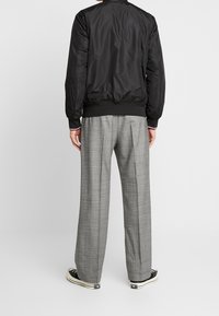 Sort bomberjakke med elastiske manchetter, parret med lysegrå ternede bukser med vidde. Model iført sorte sneakers.