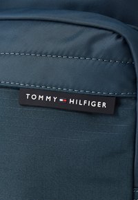 Tamnoplava tkanina ima teksturiranu površinu, uz istaknutu crnu oznaku s natpisom "TOMMY HILFIGER" u crvenim i bijelim akcentima.