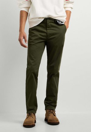 Mann trägt olivgrüne Slim-Fit-Hose, braune Schnürstiefel und ein weißes Langarmhemd mit hochgekrempelten Ärmeln.