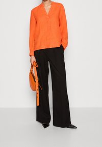 Haut en lin orange avec un col en V, manches longues, associé à un pantalon noir à jambes larges et un sac à main orange avec une longue bandoulière. Chaussures noires à bout pointu.