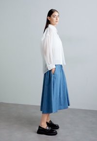Hvid gennemsigtig bluse, blå denimnederdel med lommer, sorte platform sko. Modellen står mod en lys grå baggrund. Strukturerede stoffer.