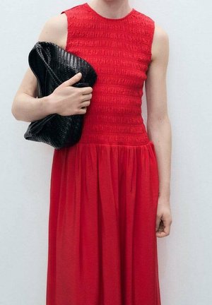 Kobieta w czerwonej sukience bez rękawów trzyma dużą czarną teksturowaną kopertówkę na białym tle.