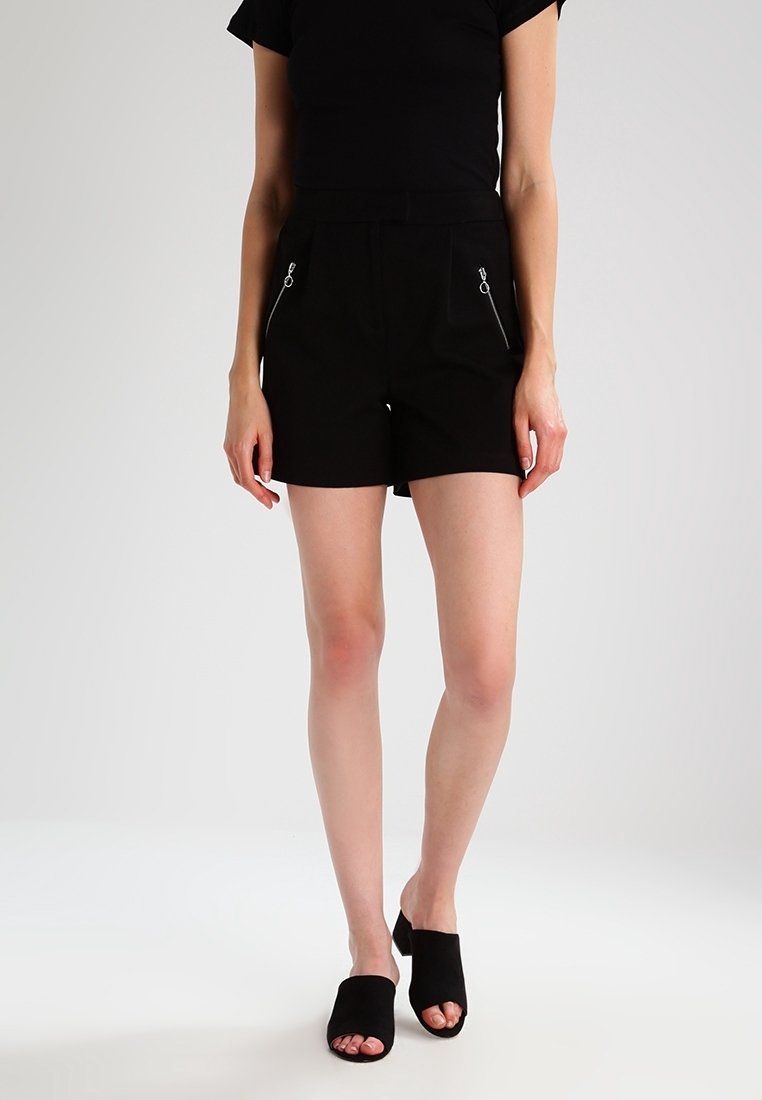 Femme portant un short taille haute noir avec des poches à fermeture éclair, un haut noir à manches courtes et des mules noires à bout ouvert sur fond blanc.