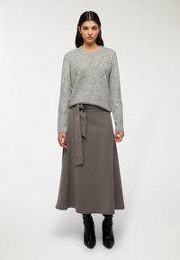 Donna che indossa un maglione grigio con dettagli a fiocco di perle, una gonna midi grigia legata in vita e stivaletti neri con tacco, in piedi contro uno sfondo chiaro e semplice.