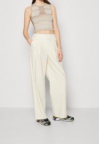 Beige ärmlös topp med ribbad textur och grafiska detaljer, i kombination med vita vida byxor och svarta sportskor med gröna accenter.
