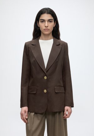 Kobieta z długimi ciemnymi włosami, ubrana w brązową marynarkę na białym topie, stojąca z neutralnym wyrazem twarzy na jednolitym tle.