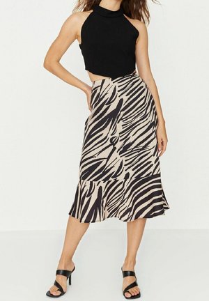 Frau posiert in einem ärmellosen schwarzen Crop-Top, einem beigen und schwarzen Zebra-Midirock und schwarzen High-Heel-Sandalen vor einem schlichten Hintergrund.