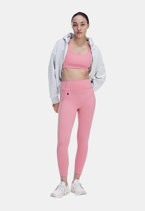Frau steht, trägt pinkes Sport-BH und Leggings, grauen Kapuzenpullover mit Reißverschluss und weiße Laufschuhe, vor weißem Hintergrund.