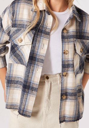 Kobieta nosząca beżową, granatową i niebieską koszulę w kratę na guziki, na białym t-shircie i kremowych spodniach.