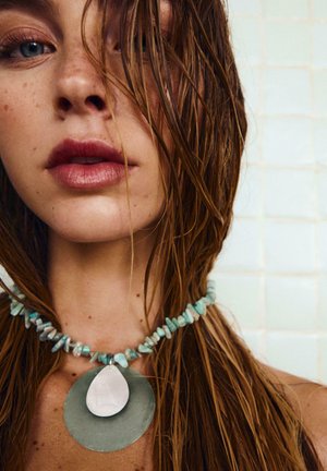 Közeli kép egy nedves hajú nőről, aki kékeszöld kőből készült nyakláncot visel nagy, csepp alakú medállal.