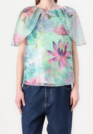 Personne portant un haut transparent à motif floral avec de grandes fleurs pastel et des manches en forme de cape, associé à un jean en denim bleu foncé.