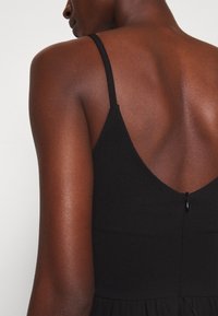 Gros plan d'une personne portant une robe noire sans manches avec de fines bretelles, montrant le haut de son dos et son épaule sur un fond uni.
