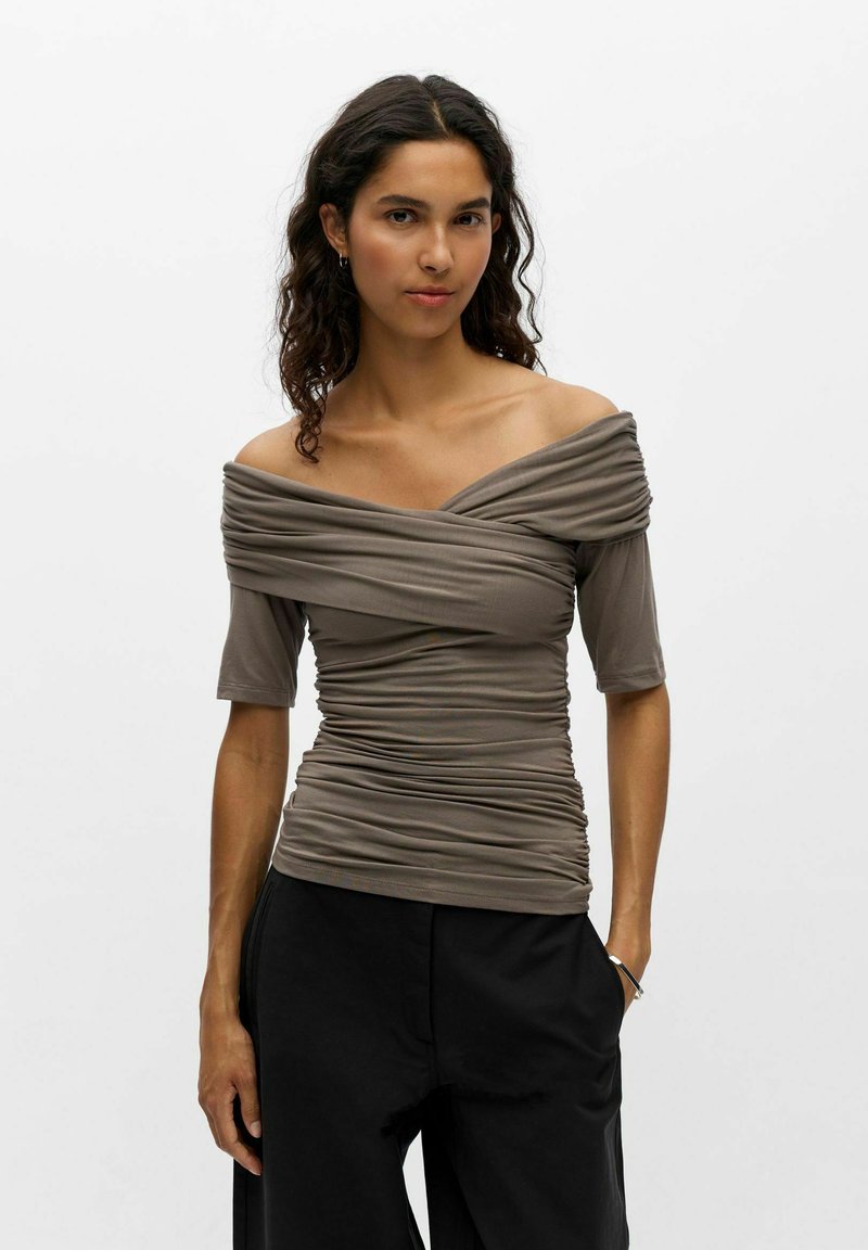 Femme aux cheveux bouclés foncés portant un haut taupe à épaules dénudées froncé et un pantalon noir, debout avec une main dans la poche sur fond blanc.