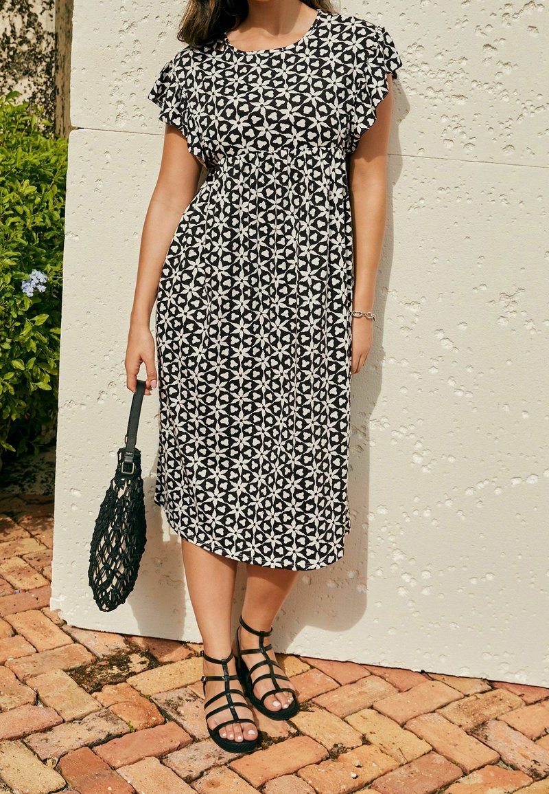 Femme portant une robe midi à motifs noirs et blancs, des sandales noires à lanières, et tenant un sac à main tressé noir, debout sur un sol pavé en briques.