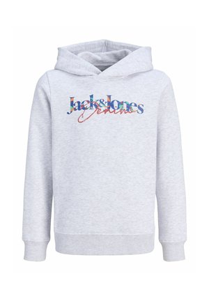 Junge Person steht, trägt einen hellgrauen Hoodie mit buntem Text, weite schwarze Jeans und graue Sneakers vor einfachem Hintergrund.