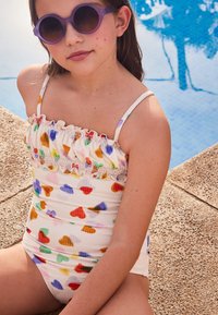 Λευκό ολόσωμο μαγιό με πολύχρωμα σχέδια καρδιών, λουλούδια στον λαιμό και λεπτές τιράντες, συνδυασμένο με στρογγυλά μωβ γυαλιά ηλίου.