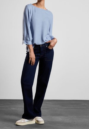 Blusa azzurra chiara a maniche lunghe con polsini arricciati, abbinata a jeans dritti blu scuro e sneakers beige e bianche. Tessuto morbido.