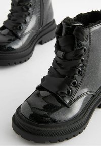 Bottes de combat en cuir verni noir avec une finition pailletée, semelle en caoutchouc épaisse et lacets en satin. Doublées d'un matériau noir doux pour le confort.