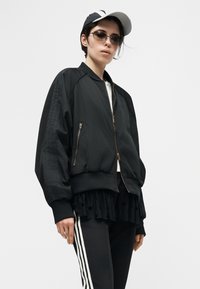 Άτομο που φοράει υπερμεγέθη μαύρο μπουφάν bomber, μαύρο παντελόνι με λευκές ρίγες στα πλαϊνά, στρώση φούστας με μαύρα πουά, λευκή καπέλο και γυαλιά ηλίου.