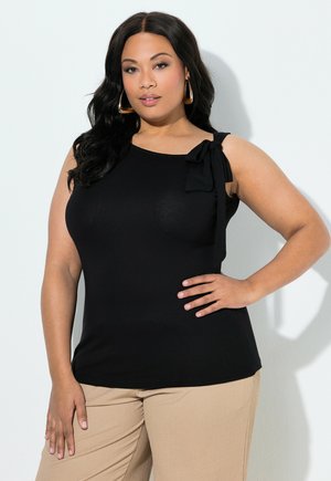 Femme aux longs cheveux noirs portant des boucles d'oreilles dorées en créole, un haut noir sans manches avec un nœud sur l'épaule, et un pantalon beige, posant avec une main sur la hanche.