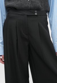 Melnas kroku bikses ar gludu faktūru, tām ir plata jostasvieta un divas tumšas pogas. kombinētas ar gaiszili svītrainu kreklu.