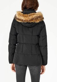 Doudoune noire avec capuche doublée de fourrure. Présente un design matelassé, des manches longues et une coupe ajustée. La bordure en fourrure est de couleur marron clair.