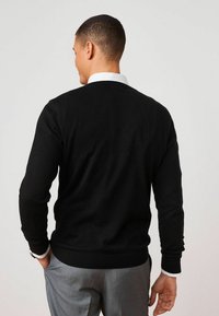 Suéter negro de punto con cuello redondo, mangas ajustadas y dobladillo acanalado. Llevado sobre una camisa blanca con cuello, destacando un aspecto limpio.