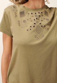 T-shirt vert olive à manches courtes avec broderie florale et découpes autour de l'encolure. Texture de tissu douce et lisse.