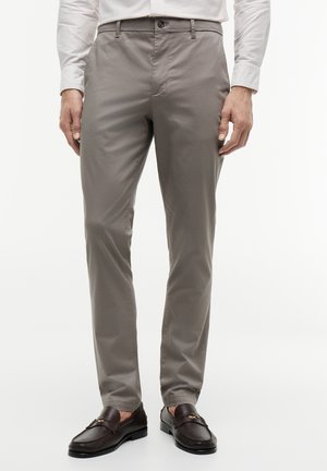Man draagt taupekleurige slim-fit broek gecombineerd met donkerbruine leren loafers en een licht gestreept overhemd met de mouwen naar beneden.