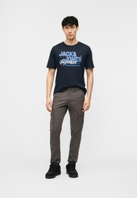 Jaunas vyras stovintis, dėvintis tamsiai mėlyną Jack & Jones grafikinį marškinėlį, pilkas krovinių stiliaus kelnes ir juodus sportbačius priešais paprastą baltą foną.