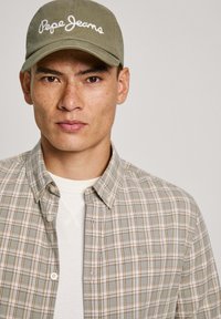 Cappello in olive con ricamo "Pepe Jeans", camicia a quadri beige e teal, tessuto testurizzato, colletto con bottoni, indossata sopra una camicia bianca.