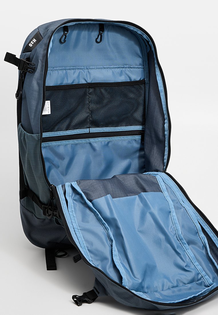 Blauer und grauer Rucksack, der geöffnet ist und mehrere Innenfächer zeigt, darunter Netzfächer und Reißverschlussabschnitte.