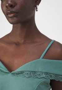 Rochie teal cu design off-shoulder, prezentând dantelă la decolteu și bretele subțiri. Material neted cu textură moale. Cercei cercuri argintii.