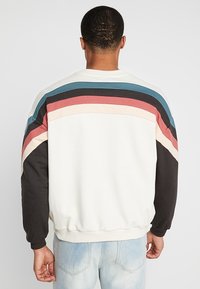 Man som bär en vit sweatshirt med blå, svart, röd och krämfärgade ränder över axlarna och ärmarna, tillsammans med ljusblå jeans.