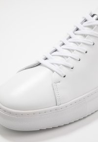 Bílé kožené tenisky s plochými bílými tkaničkami, zaoblenou špičkou, kontrastním šitím a texturovanou gumovou podešví. Minimalistický, hladký povrch designu.