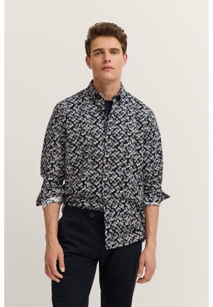 Junger Mann trägt ein marineblaues Hemd mit Blumenmuster, hochgekrempelten Ärmeln und schwarze Hose, steht vor einem einfarbigen hellen Hintergrund.