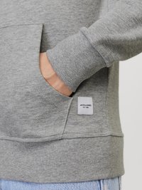Sweatshirt gris avec des poignets côtelés et une poche latérale. Présente une étiquette en tissu avec "JACK & JONES EST. 1989." Texture douce, design décontracté.