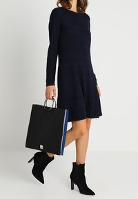 Abito blu notte a maniche lunghe in tessuto testurizzato, borsa quadrata nera con accessori in argento e dettagli blu, abbinata a stivaletti neri alla caviglia.