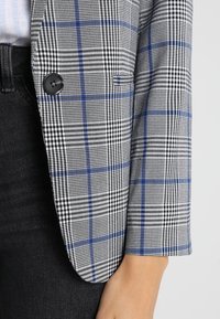 Šedý a černý kostkovaný blazer s modrými akcenty, s jedním černým knoflíkem a přizpůsobeným střihem. Měkká tkanina a strukturovaný tvar.