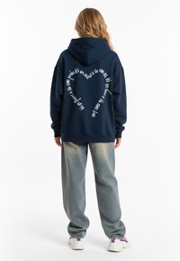 Námornícky modrá mikina s kapucňou, na chrbte s bielym textom v tvare srdca, skombinovaná so svetlošedými, zúženými nohavicami a bielymi teniskami.
