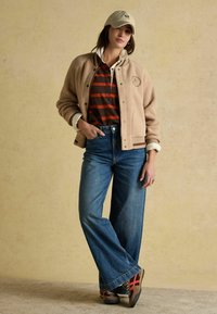 Naine kannab beeži bomber-jakki, triibulist polo särki, laia jalaga teksaseid, spordijalatsite ja beeži nokamütsi, seistes ülevaatuseta seina vastas.