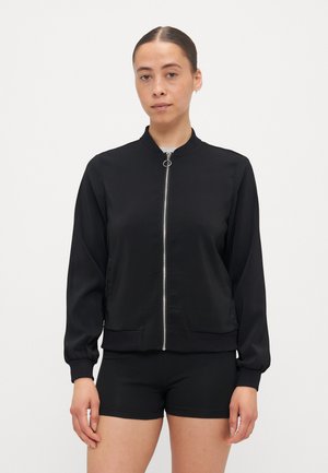 Veste noire zippée avec un col et des poignets côtelés, présentant une texture lisse et brillante ainsi que des poches latérales. Portée sur un short noir.