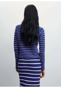 Personne aux longs cheveux noirs portant un pull et une jupe rayés horizontalement bleu et blanc, debout de dos sur un fond uni.