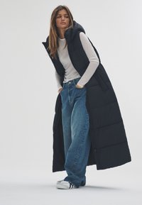 Fekete kapucnis steppelt mellény, hosszú fazon, fehér hosszú ujjú inggel és lazán szabott kék farmerrel párosítva, fehér tornacipővel kiegészítve.