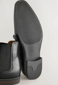 Botas negras de cuero hasta el tobillo con paneles laterales elásticos, suelas de goma texturizadas con un patrón geométrico y acentos marrones contrastantes en el interior.