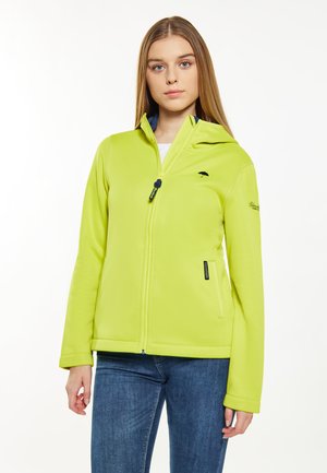 Jonge vrouw draagt een limegroene jas met rits en een klein paraplu-logo op de borst en blauwe spijkerbroek, staand tegen een witte achtergrond.