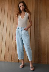 Ljusblå högmidjade jeans med en avslappnad passform, tillsammans med en krämfärgad stickad linne med djup V-ringning. Modellen har på sig texturerade sandaler.